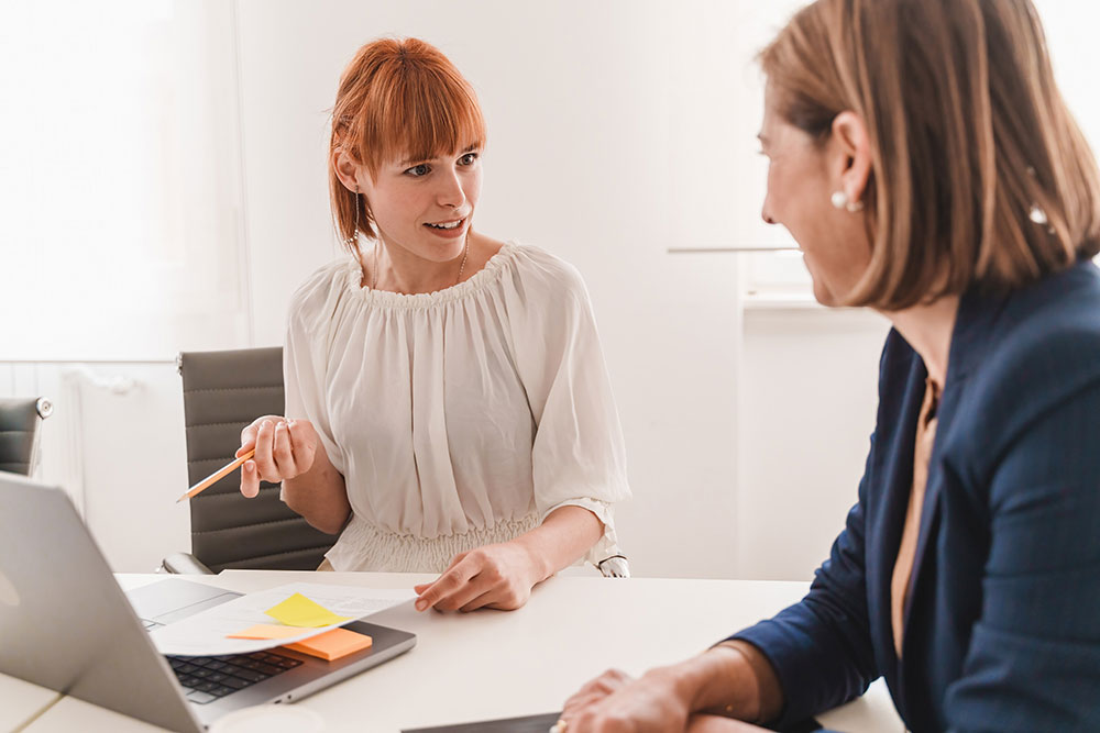 Zwei Frauen, sich gegenübersitzend, sprechen am Schreibtisch miteinander, Bildnachweis: iStock/ lomp