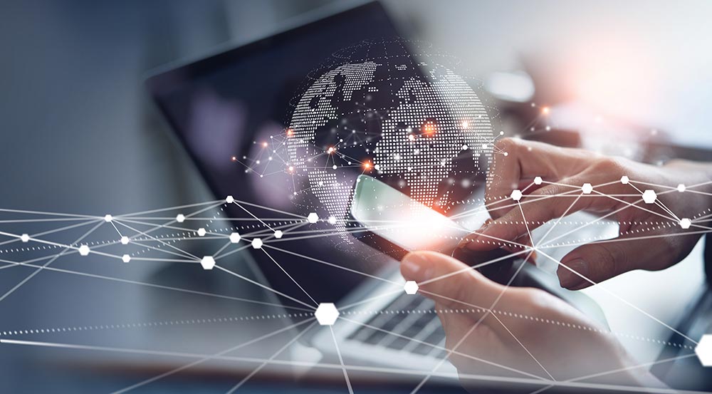 Hände halten und bedienen ein Smartphone, im Hinterund eine Weltkugel vor einem Laptop, Foto: iStock/TippaPat