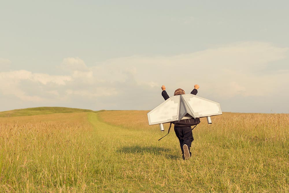 Person mit erhobenen Armen und selbst gebauten Flugzeugflügeln läuft über ein Feld, Bildnachweis: gettyImages/RichVintage