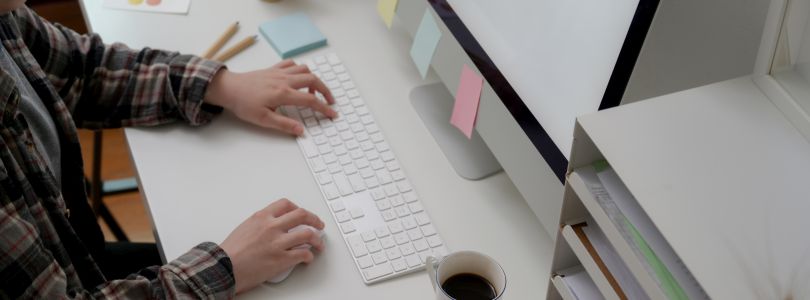 Person sitzt am Schreibtisch, darauf Kaffetasse, Bildschirm mit Notizzetteln und Aktenablage